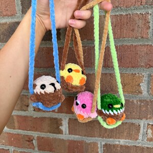 May include: Four small, plush, crocheted animal charms hanging from soft yarn loops. The charms include a brown and white otter on a blue loop, a yellow chick on a brown loop, a pink creature on a brown loop, and a green mallard duck on a lime green loop.