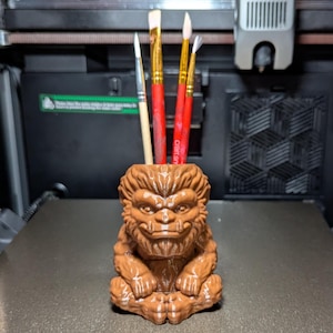 May include: A brown, 3D-printed pen holder shaped like a monster with a fierce expression. The pen holder contains several paint brushes with red and yellow handles. The pen holder is sitting on a gray surface.