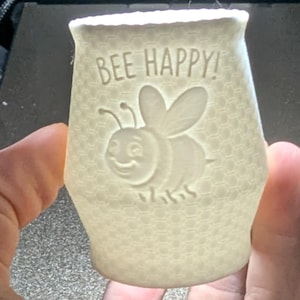 May include: A small, off-white, cylindrical container with a honeycomb pattern. The words "BEE HAPPY!" are etched above a smiling bee illustration. The container is held by a hand.