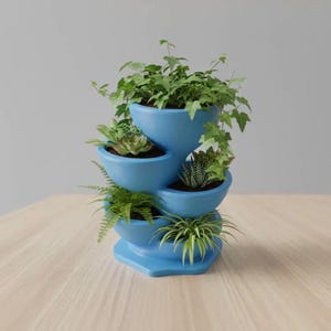 May include: A multi-tiered, blue ceramic planter filled with various green plants. The planter has a unique, cascading design with several bowl-shaped sections, each holding different types of greenery. The planter is on a light-colored surface.