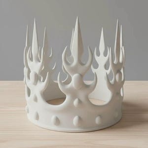 May include: A white ceramic crown with a series of tall, flame-like spikes and rounded details. The crown sits on a light-colored wooden surface, with a neutral gray background.