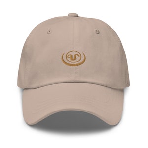 May include: A beige baseball cap with a curved brim. The cap features a gold-colored embroidered logo on the front. The cap has a six-panel design with a button on top and an adjustable strap at the back.
