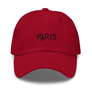 May include: A red baseball cap with the word "PARIS" embroidered in black across the front. The Eiffel Tower is depicted as the letter "A" in "PARIS". The cap has a curved brim and a fabric strap closure.