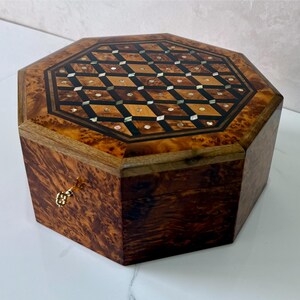 May include: Octagonal wooden jewelry box with a patterned top. The box features a burl wood exterior with a gold-colored clasp. The top is inlaid with a geometric design of dark and light wood, and mother-of-pearl accents.