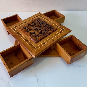 Peut inclure: Une boîte à bijoux en bois avec quatre compartiments carrés et un carré central avec un motif décoratif sculpté. La boîte est faite de bois clair et foncé avec des accents dorés.