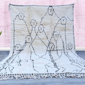 May include: A white Moroccan rug with black abstract designs. The rug has a fringe border and features a pattern of stylized faces.