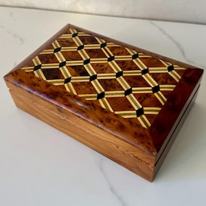 May include: A rectangular wooden box with a decorative inlaid lid. The lid features a geometric pattern of dark brown, light brown, black, and gold-colored wood. The box has a polished finish and a natural wood grain.
