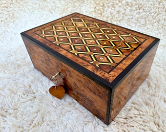 Puede incluir: Una caja de madera con un patrón decorativo incrustado en la tapa. La caja tiene una cerradura y un dije de llave en forma de corazón.