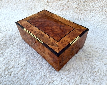 May include: A rectangular wooden box with a glossy finish. The box features a burl wood pattern on the top and sides, with dark brown and gold accents. The box has a small keyhole and a hinged lid.