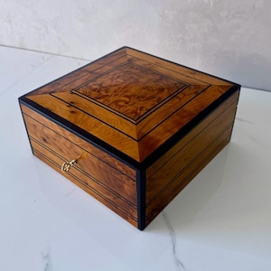 May include: A square wooden box with a decorative inlaid design on the lid. The box is made of light and dark brown wood with black trim. A small gold-colored keyhole is visible on the front. The box is likely used for storing jewelry or other small items.