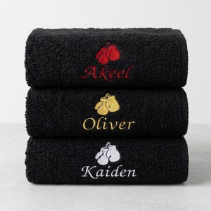 May include: A stack of three black towels, each featuring an embroidered pair of boxing gloves and a name. The top towel has red gloves and the name Akeel, the middle has gold gloves and Oliver, and the bottom has white gloves and Kaiden.