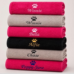 May include: A stack of six folded towels in shades of hot pink, beige, and black. Each towel features an embroidered paw print icon above a personalised name, including Winnie, Bella, Alfie, Chase, and Poppy-June, stitched in contrasting thread colours.