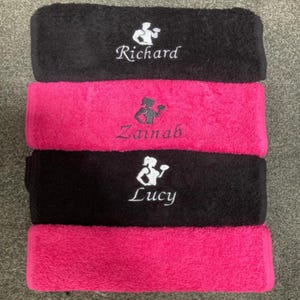 May include: A stack of four rectangular towels in alternating black and hot pink colors. The top black towel features white embroidery of a person lifting a dumbbell and the name Richard. The second pink towel has black embroidery of a person lifting a dumbbell and the name Zainab. The third black towel has white embroidery of a person lifting a dumbbell and the name Lucy.
