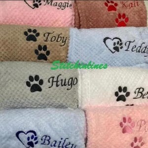 May include: A collection of soft, textured blankets in various colors including pink, brown, light blue, gray, and white. Each blanket features embroidered names such as Maggie, Kali, Toby, Teddy, Hugo, Bella, Bailey, and Poppy with paw prints.
