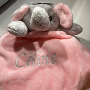 Puede incluir: Una manta de beb&eacute; de felpa con una cabeza de elefante adjunta. El elefante es gris con orejas rosas. La manta es rosa y est&aacute; bordada con el nombre "C&eacute;line" en hilo plateado. Una etiqueta est&aacute; adjunta en la parte superior.