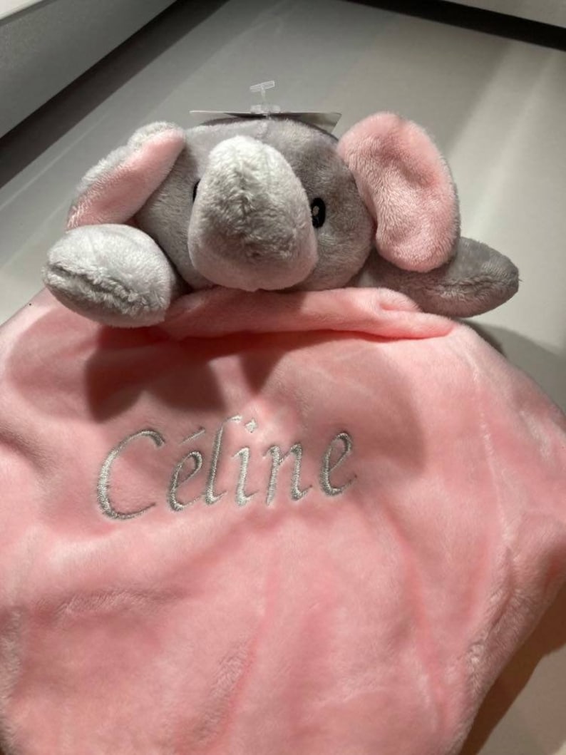 Puede incluir: Una manta de beb&eacute; de felpa con una cabeza de elefante adjunta. El elefante es gris con orejas rosas. La manta es rosa y est&aacute; bordada con el nombre "C&eacute;line" en hilo plateado. Una etiqueta est&aacute; adjunta en la parte superior.