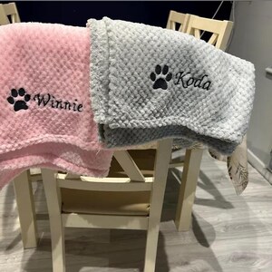 May include: Two soft, textured blankets with a waffle-like pattern. One blanket is light pink with a black embroidered paw print and the name Winnie. The other blanket is light gray with a black embroidered paw print and the name Koda.