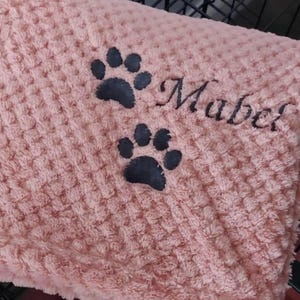 May include: A soft, light pink waffle-textured blanket featuring two dark gray embroidered paw prints and the name Mabel stitched in a cursive font. The fabric has a raised, grid-like pattern throughout the entire surface of the material.