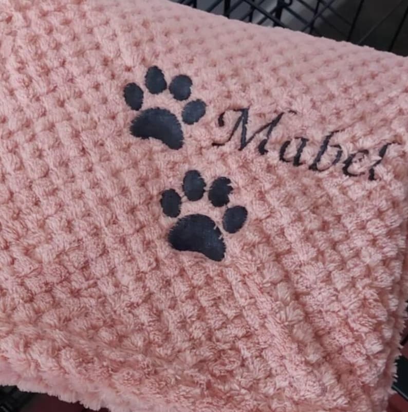 May include: A soft, light pink waffle-textured blanket featuring two dark gray embroidered paw prints and the name Mabel stitched in a cursive font. The fabric has a raised, grid-like pattern throughout the entire surface of the material.