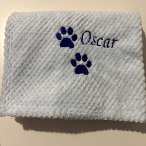 May include: A light blue textured blanket featuring two dark blue embroidered paw prints and the name Oscar stitched in dark blue thread. The fabric has a raised, waffle-like pattern across the entire surface of the folded material.
