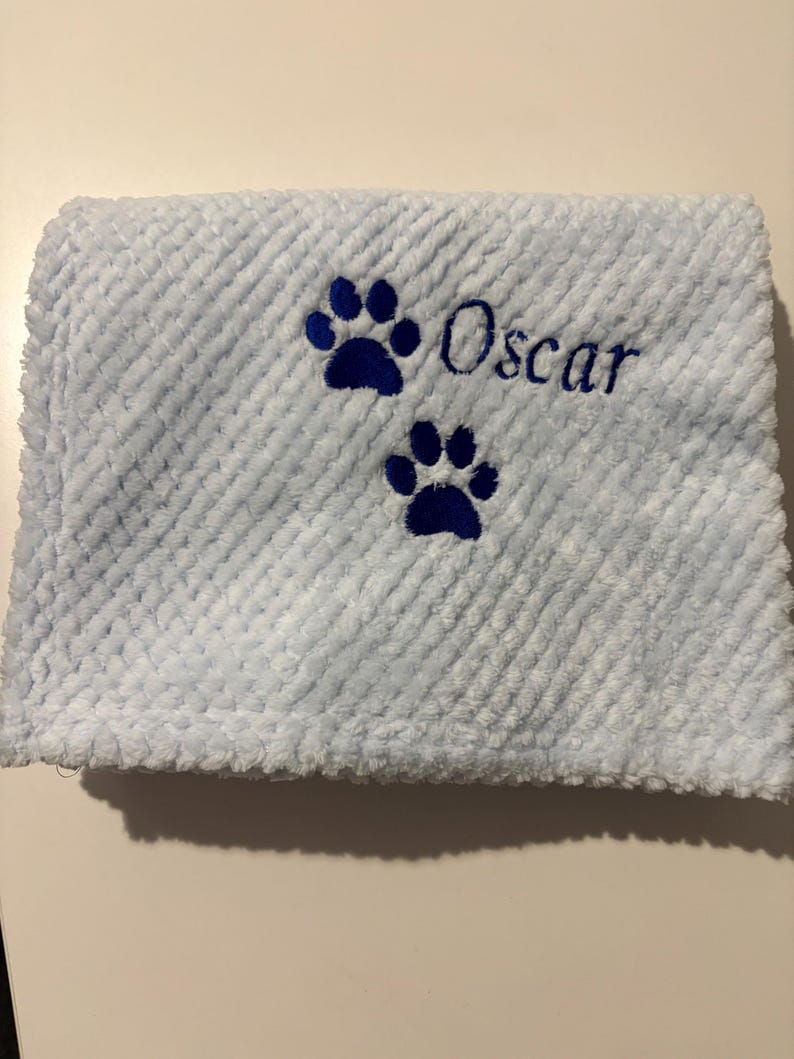 May include: A light blue textured blanket featuring two dark blue embroidered paw prints and the name Oscar stitched in dark blue thread. The fabric has a raised, waffle-like pattern across the entire surface of the folded material.