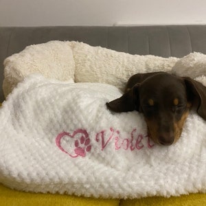 May include: A white plush pet bed with a textured, fuzzy surface. The front features pink embroidery of a heart containing a paw print next to the name Violet. A small, dark brown dachshund is resting its head on the soft, cushioned edge.