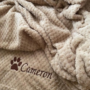 May include: A beige, textured, plush throw blanket featuring a dark brown embroidered paw print and the name Cameron stitched in a cursive font. The fabric has a soft, waffle-like pattern with deep folds and ripples throughout the material.