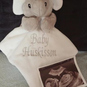 Puede incluir: Un peluche de elefante gris con una manta blanca bordada con las palabras "Baby Huskisson". Una imagen de ultrasonido est&aacute; al lado de la manta. El juguete tiene orejas, trompa y patas grises.