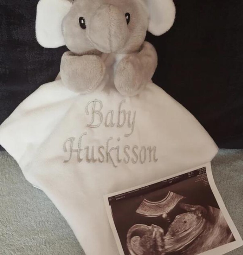 Puede incluir: Un peluche de elefante gris con una manta blanca bordada con las palabras "Baby Huskisson". Una imagen de ultrasonido est&aacute; al lado de la manta. El juguete tiene orejas, trompa y patas grises.