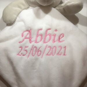 Puede incluir: Una manta de seguridad de elefante de peluche blanco con cabeza y brazos grises. La manta tiene el nombre "Abbie" y la fecha "25/06/2021" bordados en hilo rosa. El elefante tiene ojos negros.
