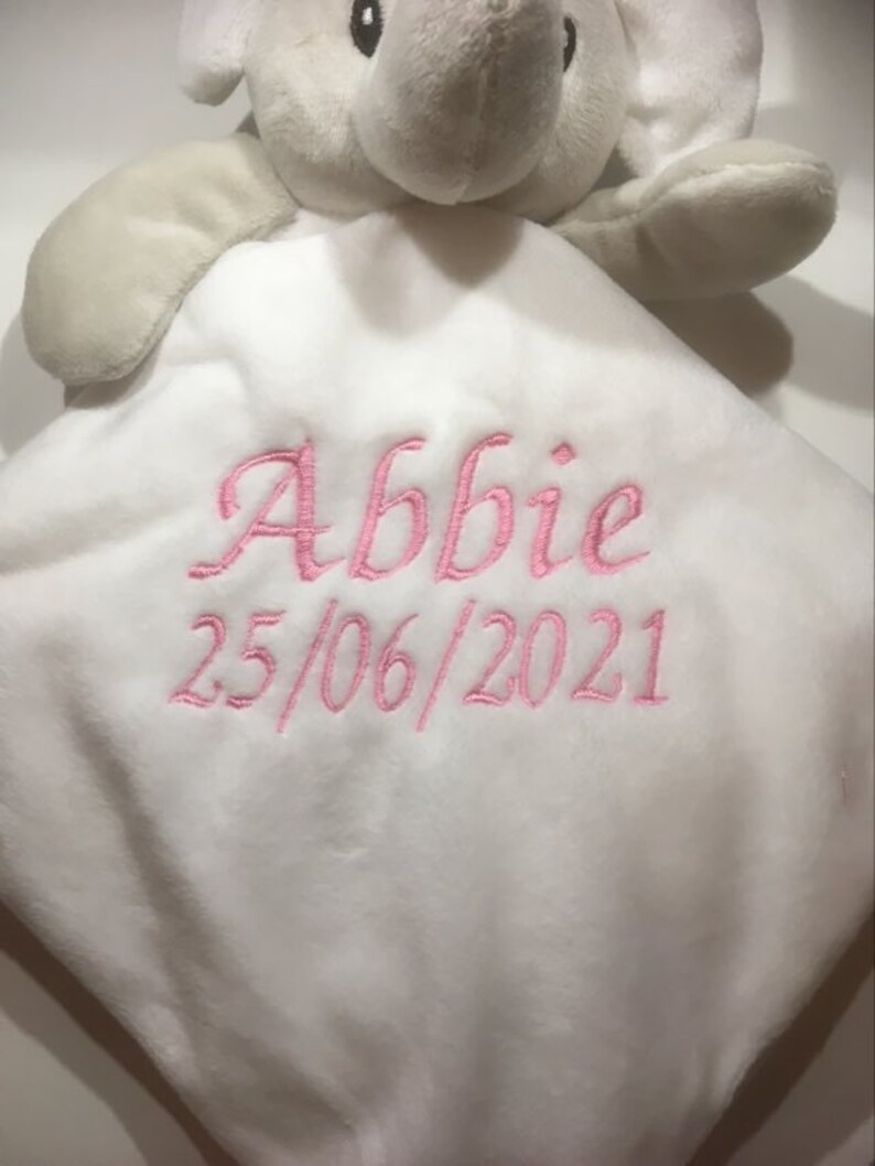 Puede incluir: Una manta de seguridad de elefante de peluche blanco con cabeza y brazos grises. La manta tiene el nombre "Abbie" y la fecha "25/06/2021" bordados en hilo rosa. El elefante tiene ojos negros.