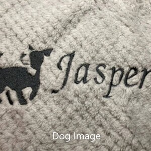 May include: A light gray textured fabric featuring black embroidery of a small dog silhouette standing next to the name Jasper written in a cursive script font. The embroidery is centered on the soft, plush material surface.