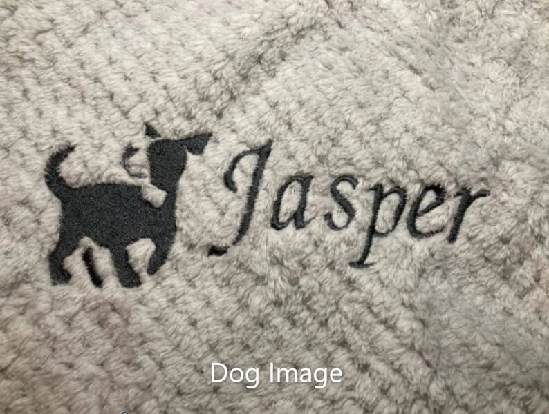 May include: A light gray textured fabric featuring black embroidery of a small dog silhouette standing next to the name Jasper written in a cursive script font. The embroidery is centered on the soft, plush material surface.
