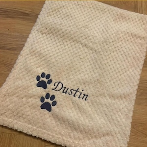 May include: A cream-colored, waffle-textured blanket featuring an embroidered design in dark blue thread. The embroidery consists of two paw prints positioned above and below the name Dustin, written in a cursive script font.