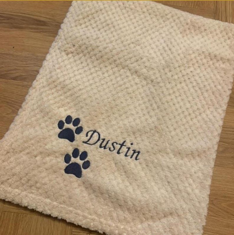 May include: A cream-colored, waffle-textured blanket featuring an embroidered design in dark blue thread. The embroidery consists of two paw prints positioned above and below the name Dustin, written in a cursive script font.