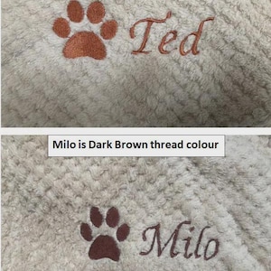 May include: A cream-colored textured fabric featuring two embroidered designs. The top design shows a bronze paw print and the name Ted. The bottom design shows a dark brown paw print and the name Milo. Text labels indicate Ted is bronze thread colour and Milo is dark brown thread colour.