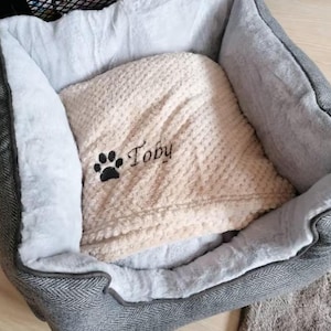 May include: A pet bed with a gray herringbone exterior and a soft white plush interior lining. Inside the bed lies a beige textured blanket embroidered with a black paw print and the name Toby in a cursive black script font.