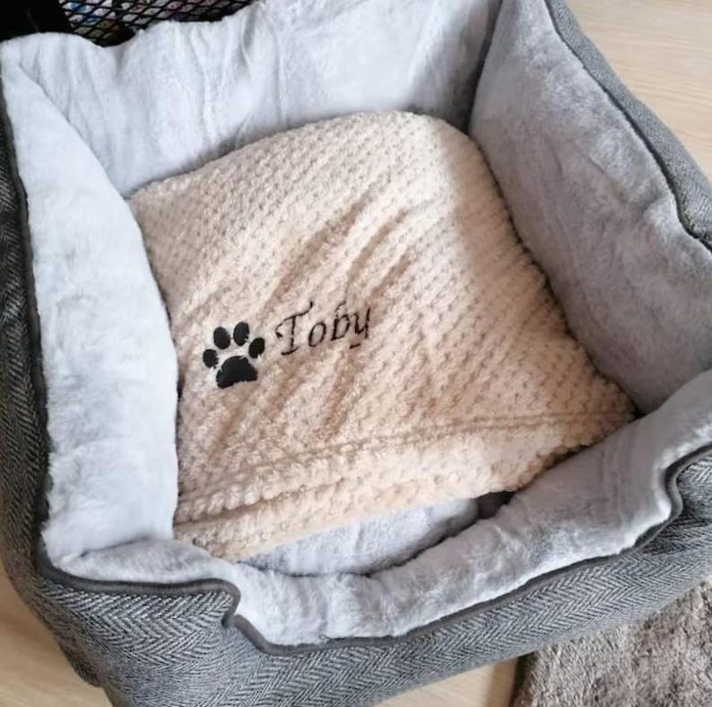 May include: A pet bed with a gray herringbone exterior and a soft white plush interior lining. Inside the bed lies a beige textured blanket embroidered with a black paw print and the name Toby in a cursive black script font.
