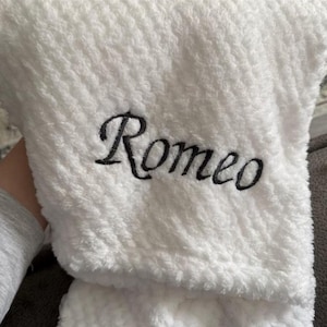 May include: A white textured plush blanket featuring the name Romeo embroidered in black script lettering. The fabric has a soft, fuzzy, and dimpled surface texture throughout, providing a tactile appearance to the personalized item.