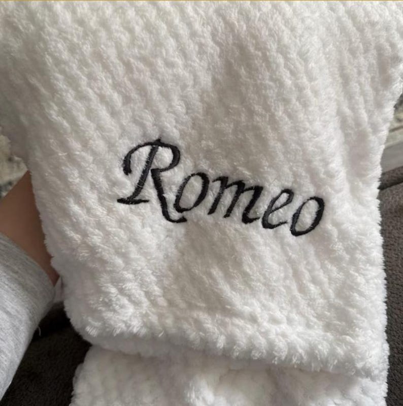 May include: A white textured plush blanket featuring the name Romeo embroidered in black script lettering. The fabric has a soft, fuzzy, and dimpled surface texture throughout, providing a tactile appearance to the personalized item.