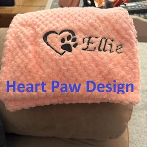 May include: A soft, light pink textured blanket featuring an embroidered black design of a heart with a paw print inside, followed by the name Ellie in a cursive script font. The fabric has a raised, bumpy, and plush surface texture.