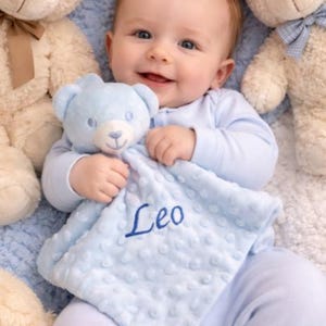 Puede incluir: Un beb&eacute; sonriente con pijama azul claro sostiene un osito de peluche azul y una manta a juego bordada con el nombre "Leo". El beb&eacute; est&aacute; rodeado de varios osos de peluche, creando una escena suave y reconfortante.