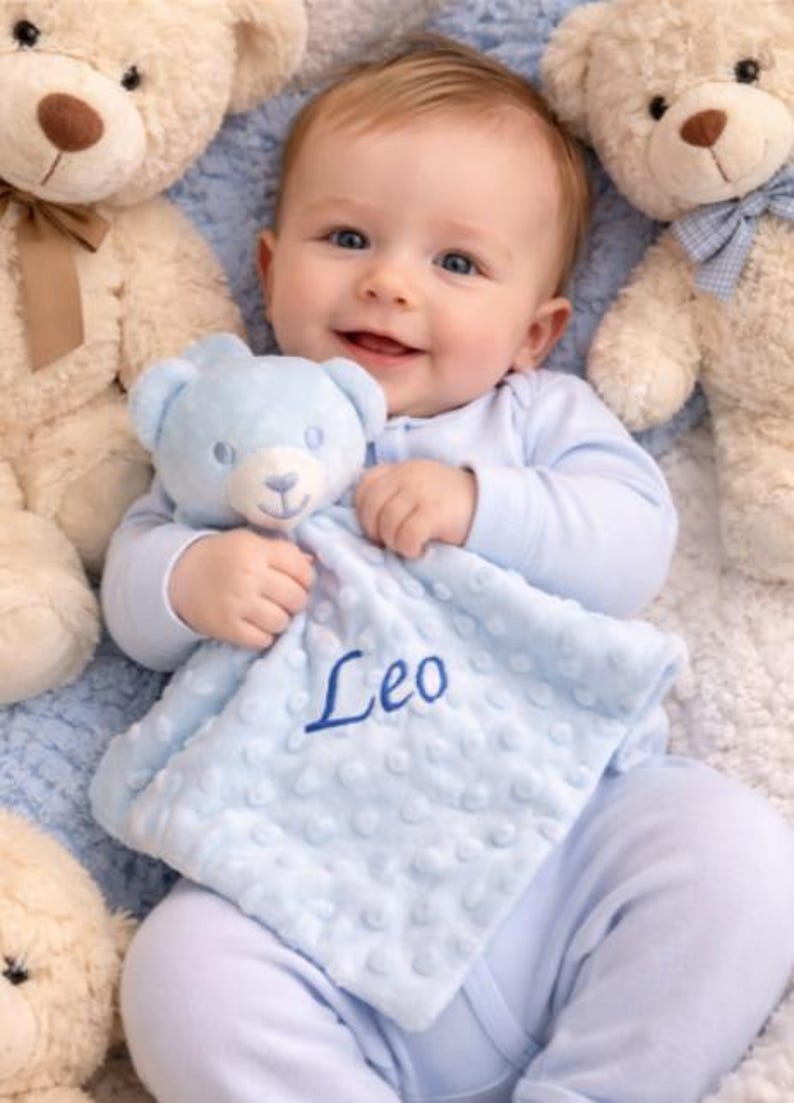 Puede incluir: Un beb&eacute; sonriente con pijama azul claro sostiene un osito de peluche azul y una manta a juego bordada con el nombre "Leo". El beb&eacute; est&aacute; rodeado de varios osos de peluche, creando una escena suave y reconfortante.