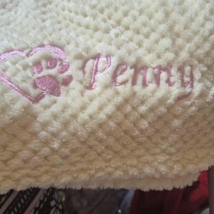 May include: A cream-colored waffle-textured blanket featuring pink embroidery. The embroidery includes a heart-shaped paw print design positioned to the left of the name Penny, which is written in a cursive script font.