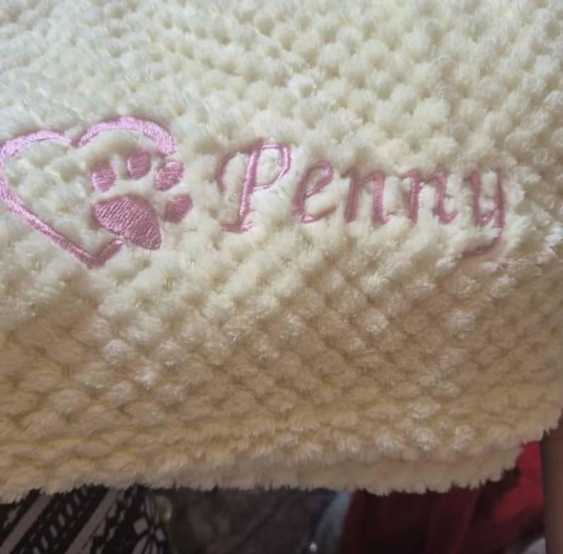 May include: A cream-colored waffle-textured blanket featuring pink embroidery. The embroidery includes a heart-shaped paw print design positioned to the left of the name Penny, which is written in a cursive script font.