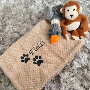 May include: A tan textured blanket featuring the embroidered name Fields and two black paw prints. Resting on the blanket are two plush toys: a gray and orange duck and a brown and cream monkey with a white belly featuring paw print details.