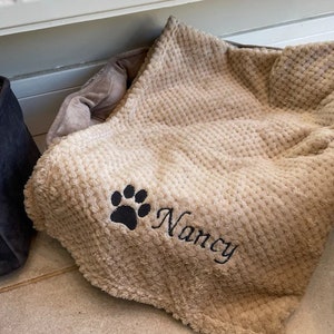 May include: A beige pet bed with a textured, plush surface. A black paw print and the name "Nancy" are embroidered on the blanket. The bed has a soft, neutral colour scheme.