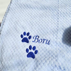 May include: A light blue waffle-textured blanket featuring two dark blue embroidered paw prints and the name Boru stitched in a dark blue cursive font. The fabric has a soft, raised grid pattern throughout the entire surface area.