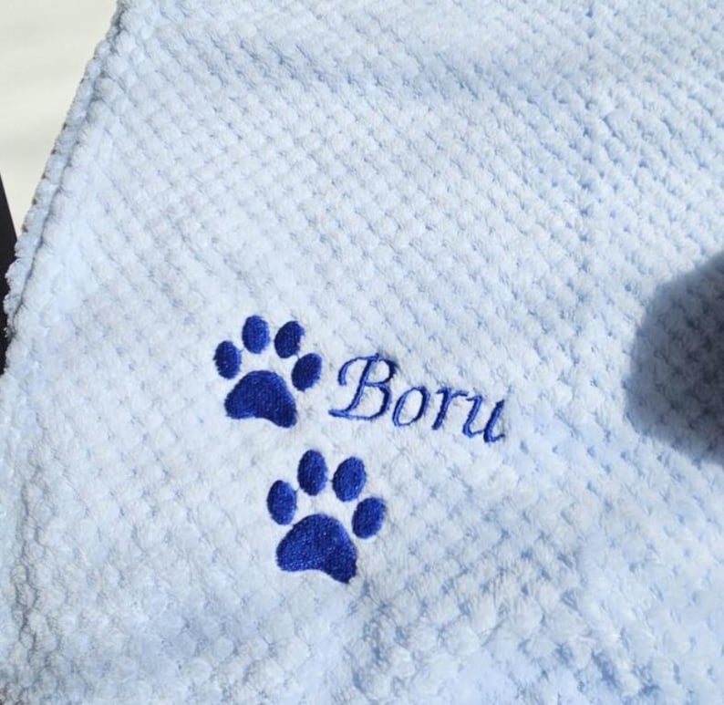 May include: A light blue waffle-textured blanket featuring two dark blue embroidered paw prints and the name Boru stitched in a dark blue cursive font. The fabric has a soft, raised grid pattern throughout the entire surface area.