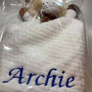 Puede incluir: Una manta de beb&eacute; blanca con el nombre "Archie" bordado en azul. Una cabeza de le&oacute;n de peluche con pelaje marr&oacute;n y beige est&aacute; adjunta. El conjunto est&aacute; empaquetado en pl&aacute;stico transparente.